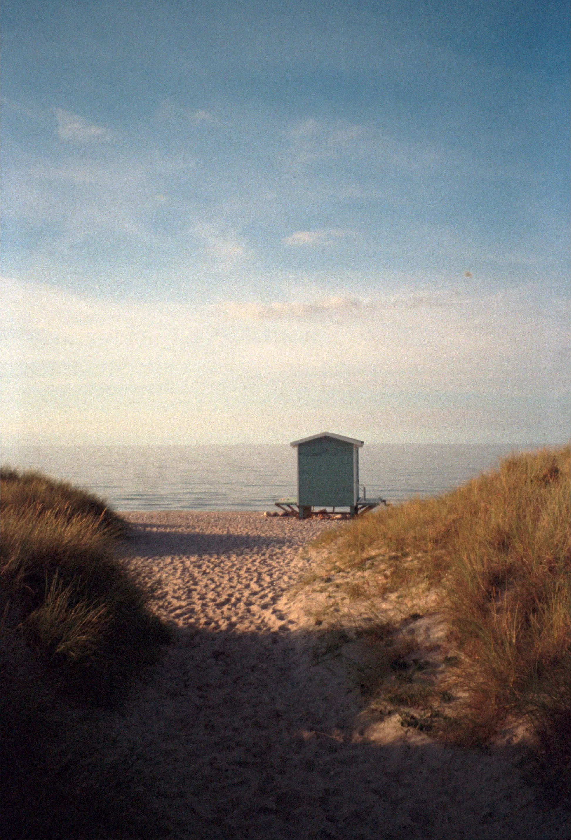 A sandy path through dark dune grass opening onto a beach, a small pale-green wooden bathing hut on stilts at the water's edge in evening light.