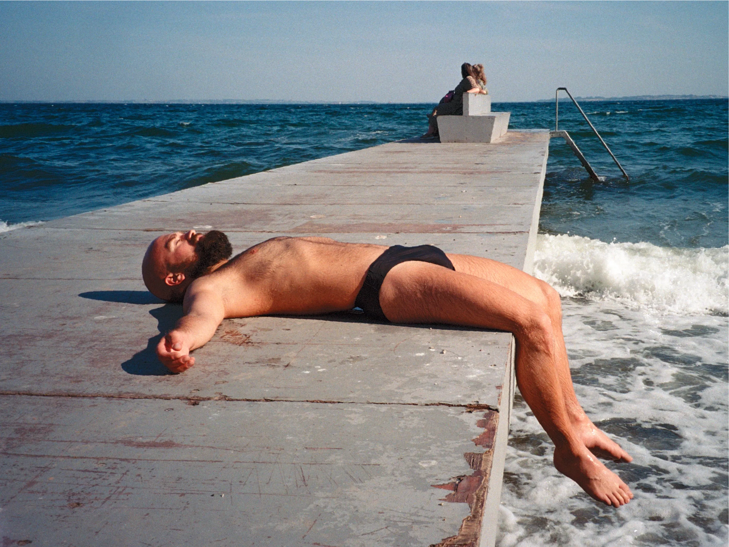 A bearded man in a black swim brief lying back on a concrete swim pier, one leg dangling toward the breaking waves; two people sit at the far end.