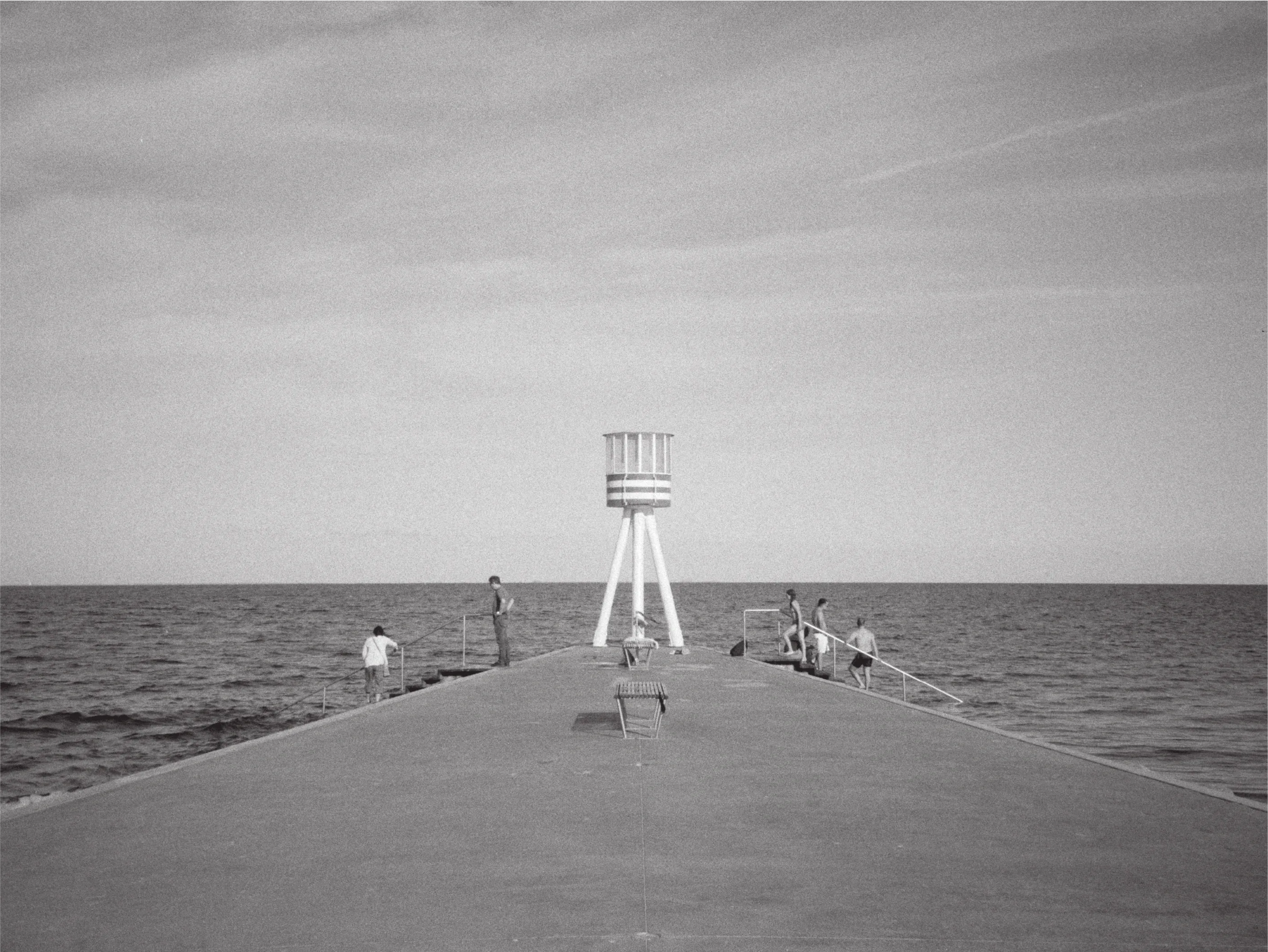 Black and white photograph of the funnel-shaped Bellevue lifeguard tower at the end of a concrete pier, people fishing and climbing from the water around it.