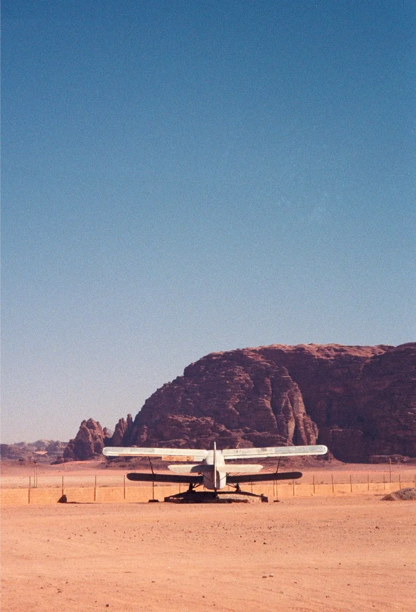 A small propeller plane parked in the desert, seen from behind, with a rock formation rising behind it.