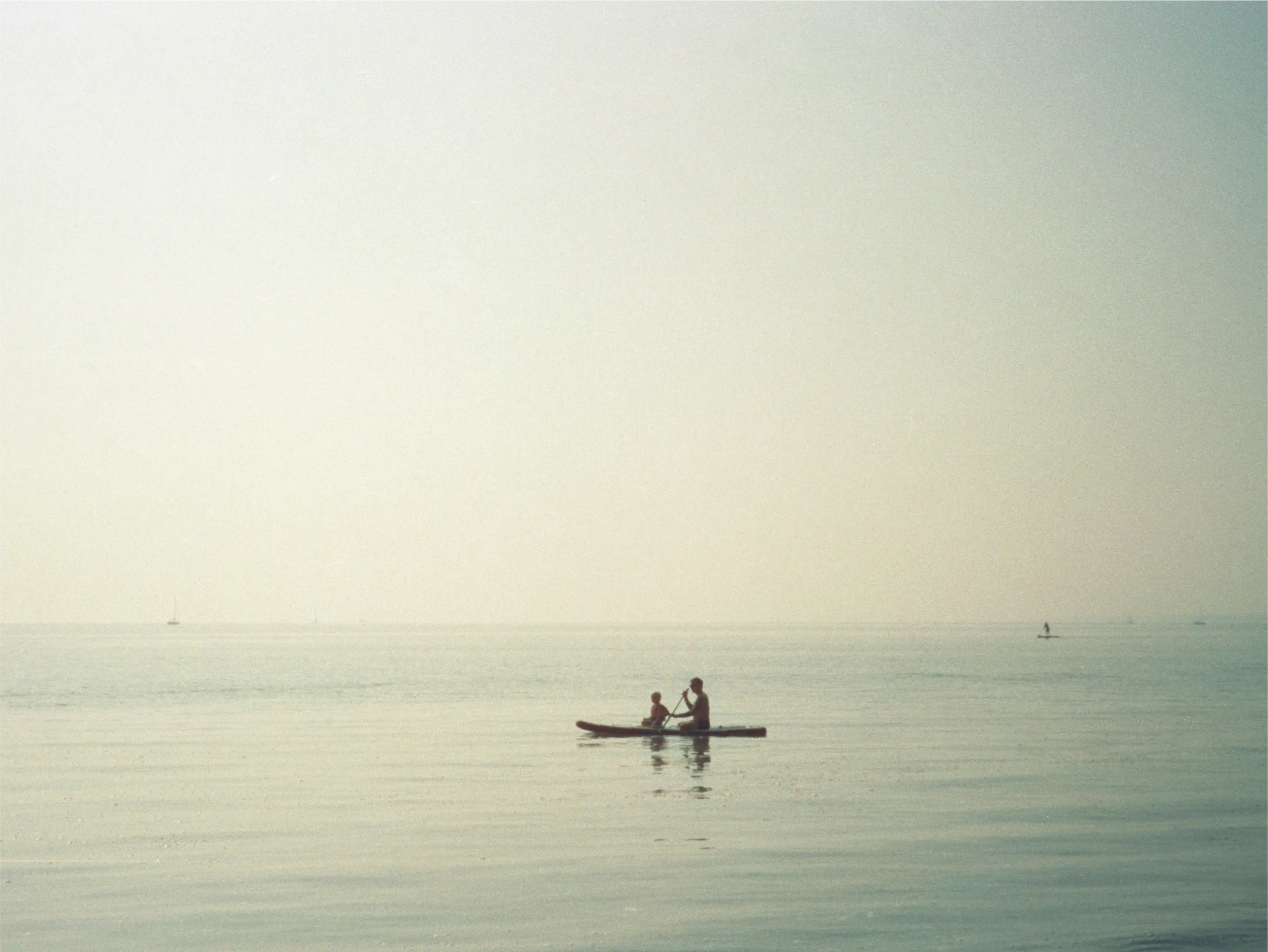 A man and a small child paddling a stand-up paddleboard across a glassy, hazy sea, distant sailboats on the horizon.