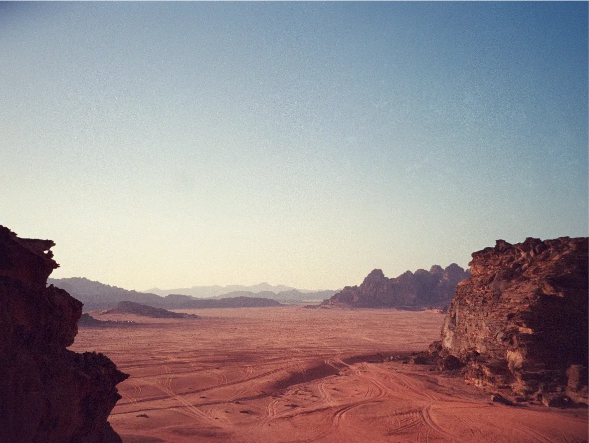 An elevated view of a red desert valley flanked by rock cliffs, tire tracks crossing the sand.