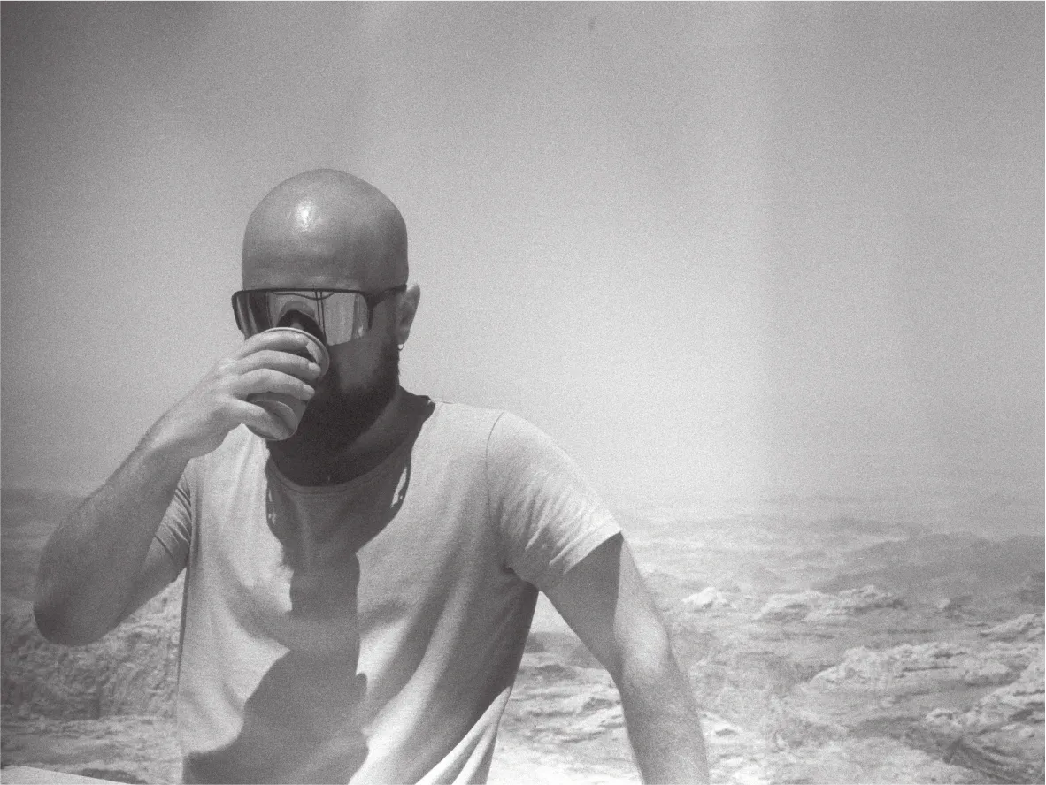 Portrait of a person drinking from a cup, sunglasses on, hazy rocky terrain behind. Black and white.