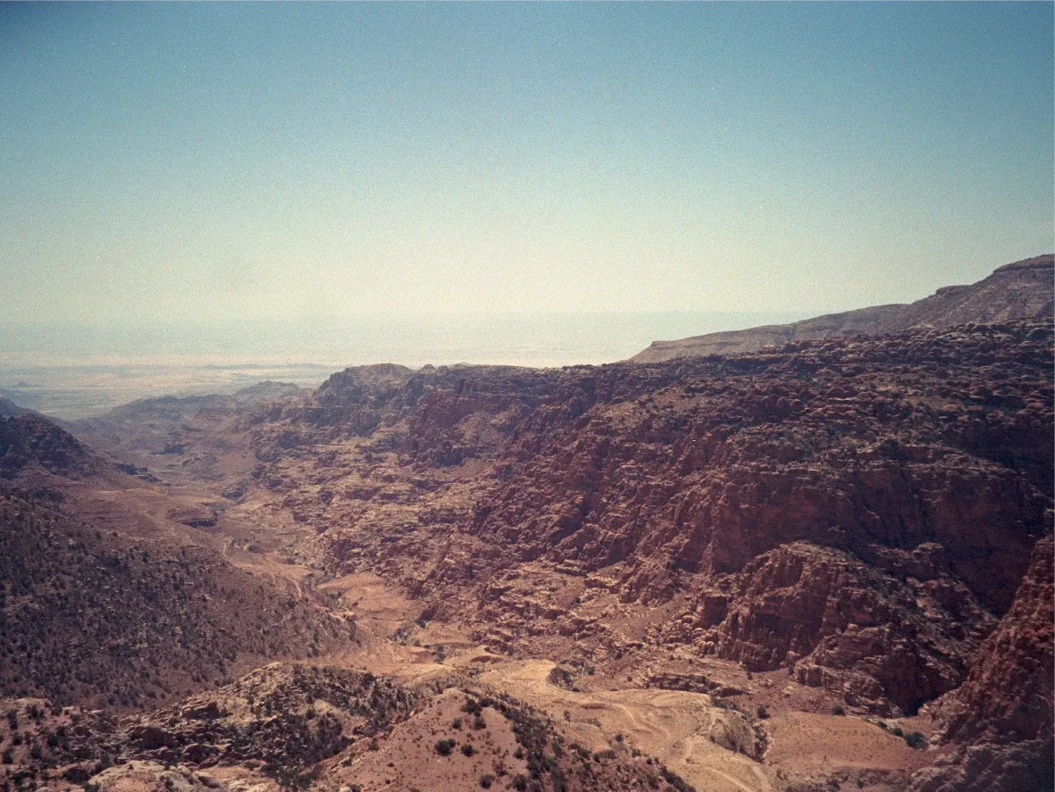 A view down into a rugged canyon from a mountainside, hazy plains in the distance.