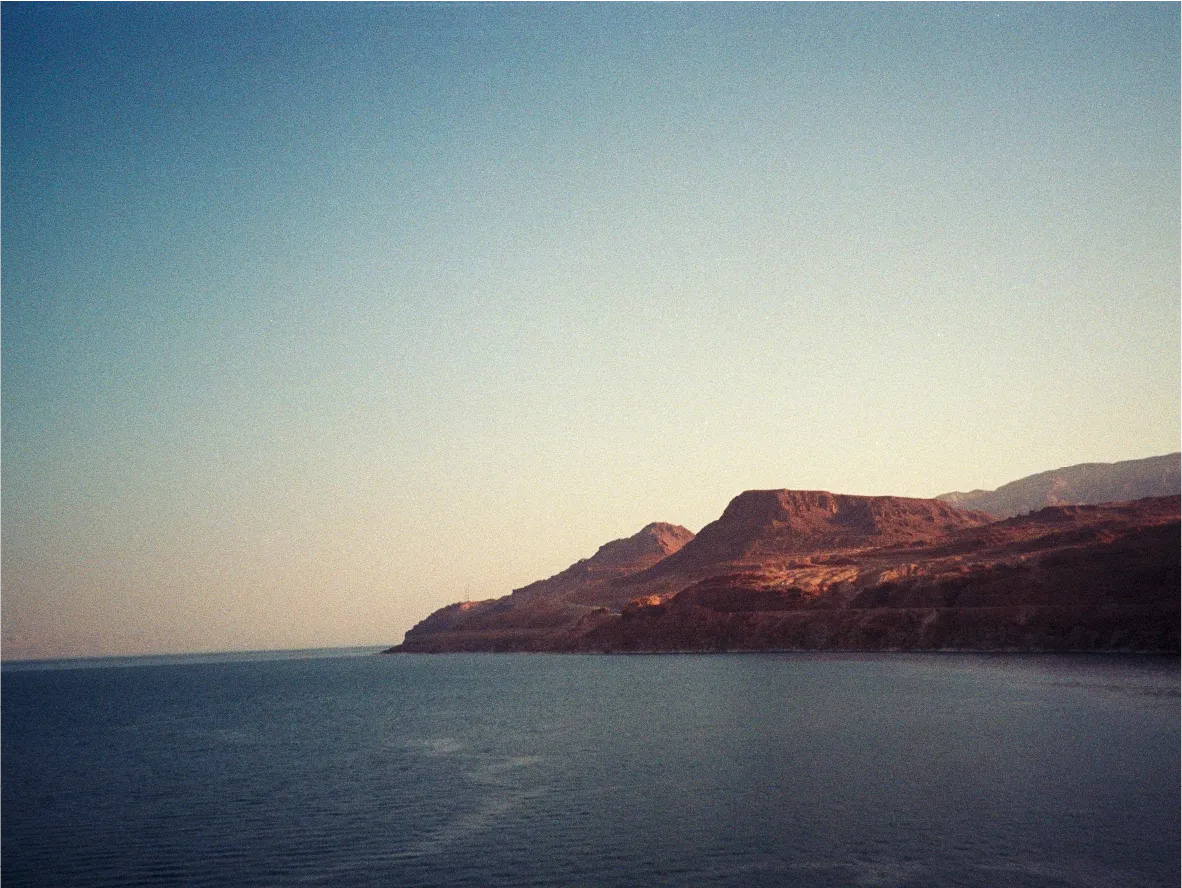 The Dead Sea coastline, calm water beneath a reddish mountainous shore.