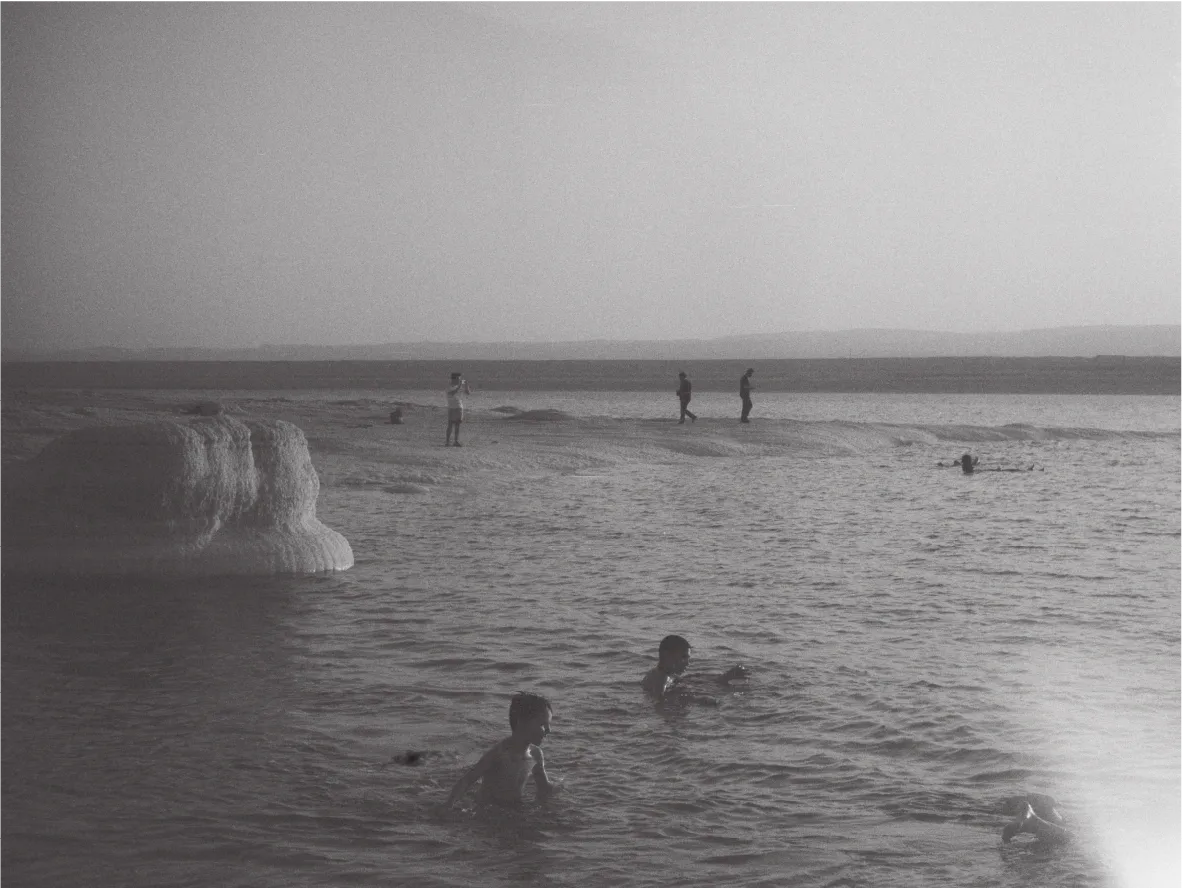 People swimming in calm water beside a white salt formation, distant shoreline. Black and white.